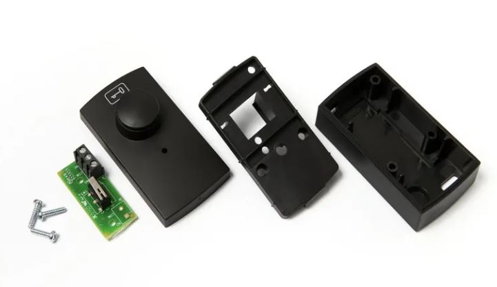 Assa Abloy - Push Button consisting of Electronic components including a black module with a knob, a green circuit board with components, and two black rectangular devices on a white background.